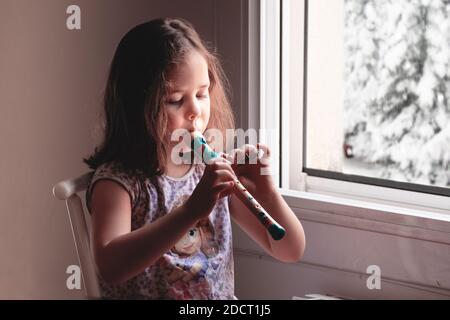 Blonde caucasian girl playing the flute next the window at home. Empty copy space for Editor`s text. Stock Photo